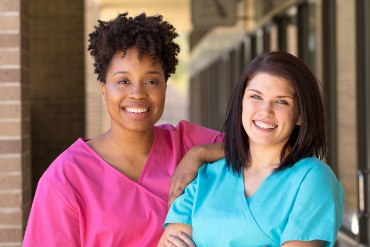 Two nurses in front of a building, wearing scrubs and smiling